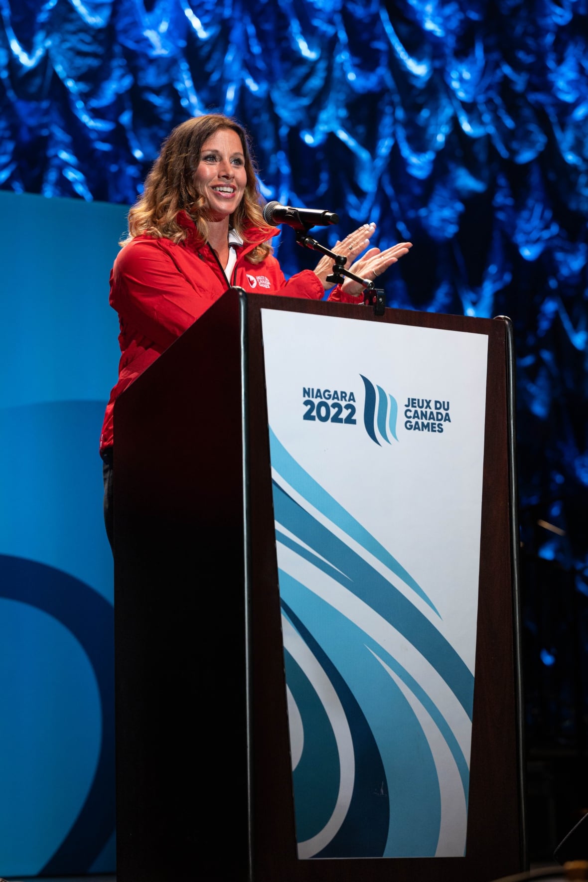 A woman wearing a red jacket stands behind a podium, with a blue background.