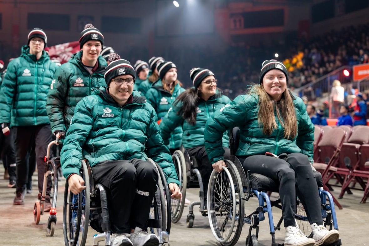 Two athletes in wheelchairs are shown in front of other athletes, all wearing matching teal coats.