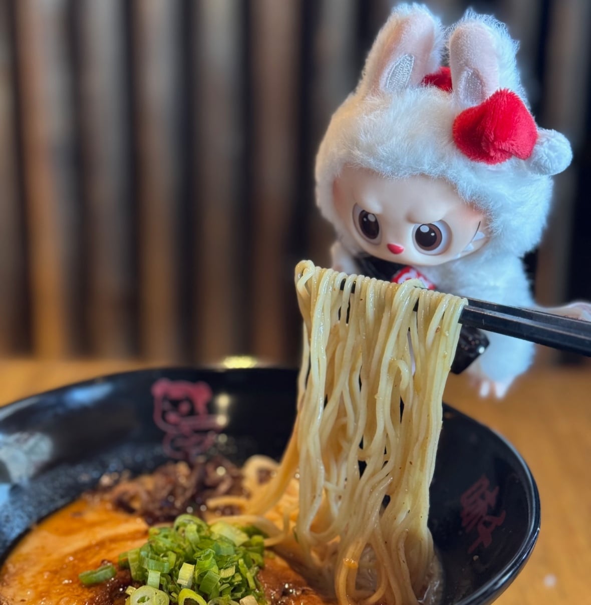 A tiny fluffy monster doll looks at a bowl of ramen, someone off campera is holding up the noodles with chopsticks