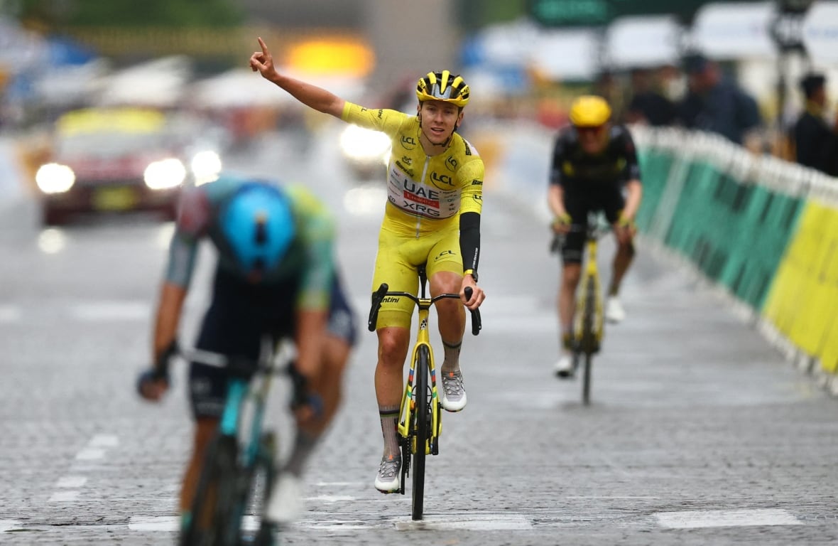 With one hand raised in triumph, a cyclist in yellow jersey rides a bike across a finish line. 