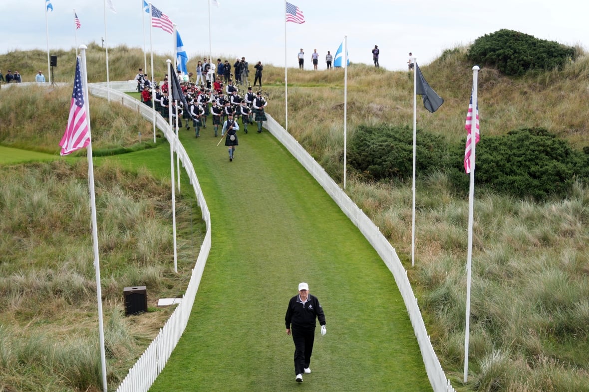 Elderly golfer walking down a grassy pathway lined with poles holding U.S. flags. Metres away, there's a marching band following behind the man.