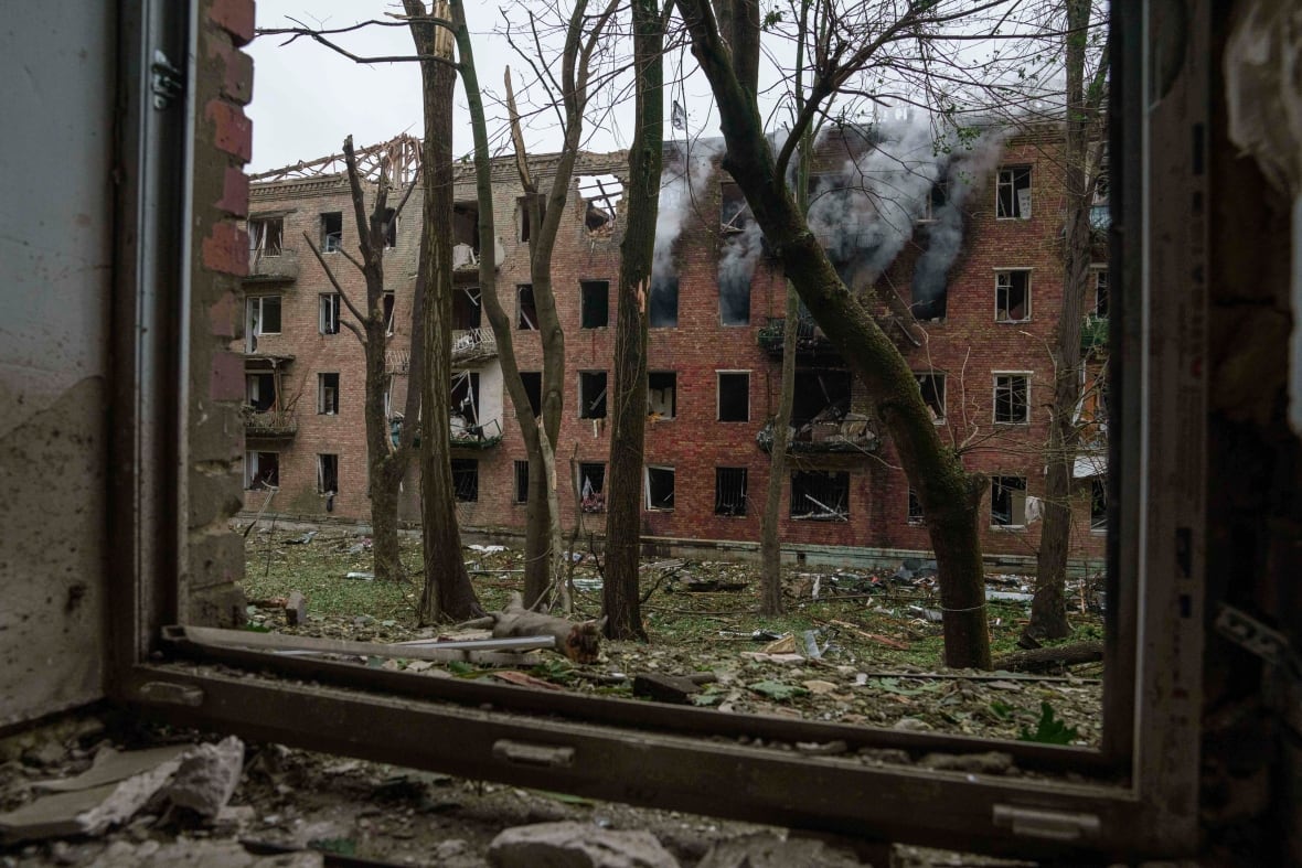 A heavily damaged low-rise building emitting plumes of black smoke from top floor windows is seen through a blown out window of another building across it.