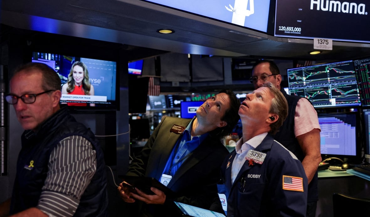 Two men look up at a screen while sitting in a busy room full of computer screens and monitors. On a screen behind them, graphs are visible. 