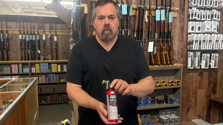 A man stands behind a counter in a gun store and holds a canister of bear spray.