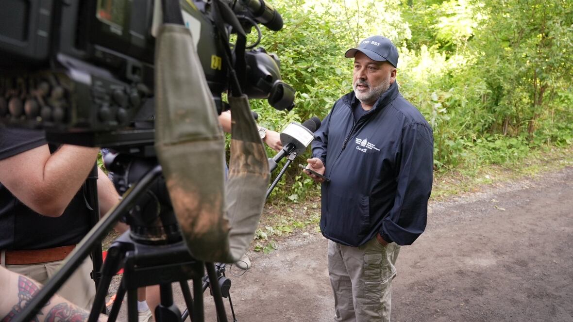A man standing in a leafy area outdoors and speaking to several cameras.