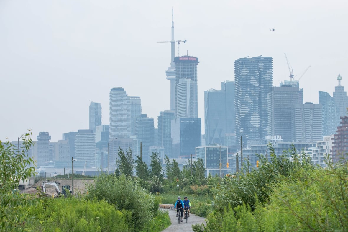 Cyclists on a path at a park surrounded by green shrubbery with a city skyline in the background.