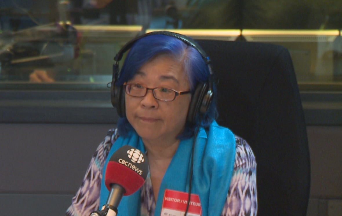 A woman with short blue hair is wearing headphone and a blue scarf and is sitting in front of a microphone.