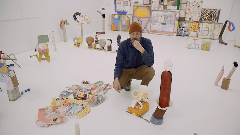 In a white room, arranged with colourful drawings and abstract sculpture, a man wearing a dark shirt and an orange toque crouches in a contemplative pose.
