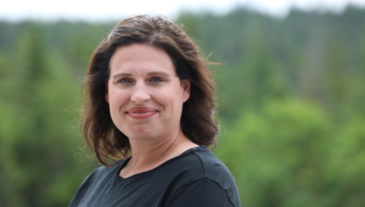 A woman with brown hair standing against a forested background.