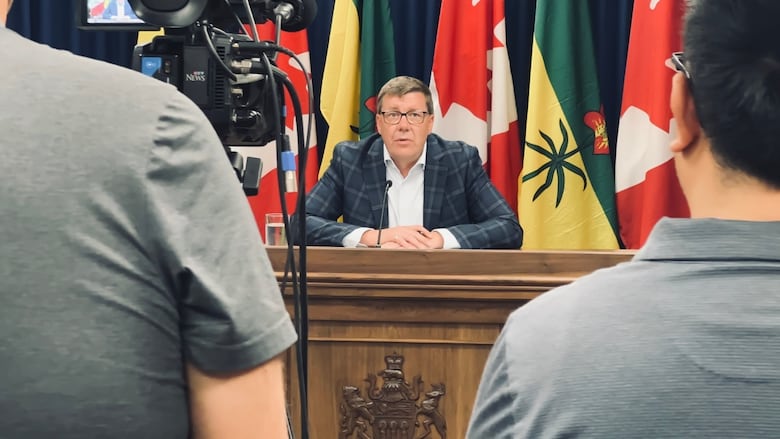 A man in a suit sits at a wooden desk, speaking before media camera.