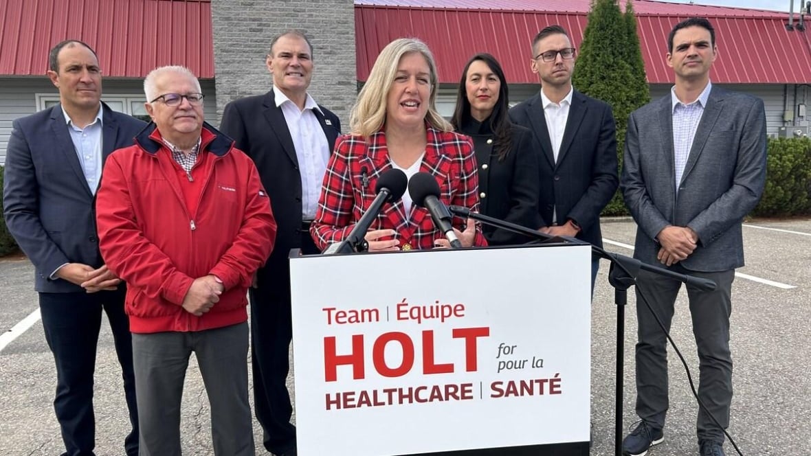 A woman with blonde hair and a red checkered blazer stands outside in front of a podium with a microphone, a group of people crowded behind her.