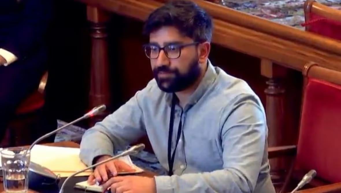 A man with brown hair and glasses wearing a blue shirt sits on a red chair at legislative assembly.