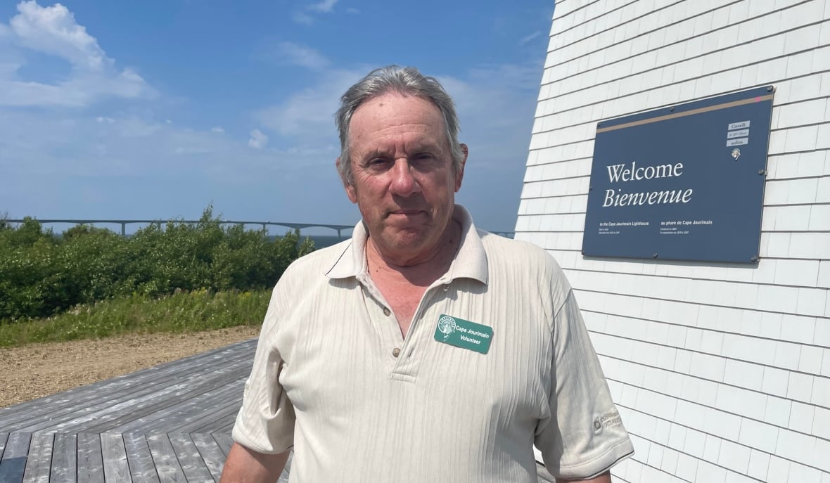 A man in a white shirt stands in front of a lighthouse with a bridge in the background.