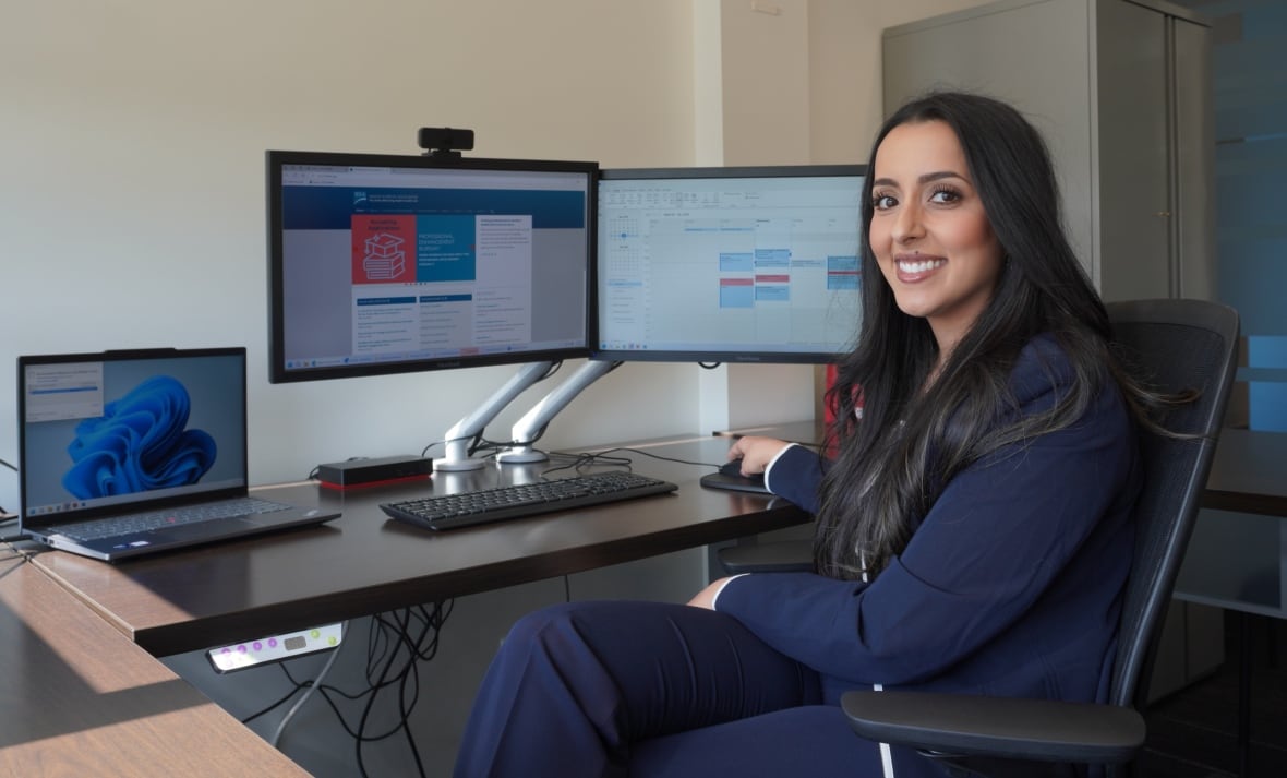 A woman with black hair in a dark blue suit sitting in front of a computer, smiling at the camera.
