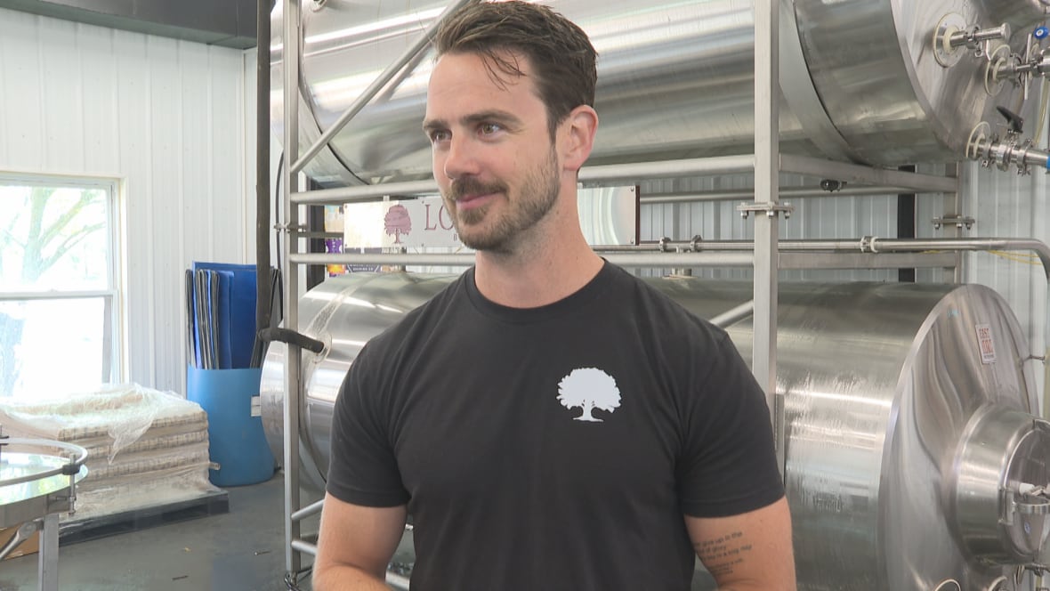 A man in a black t-shirt stands in front of brewing tanks