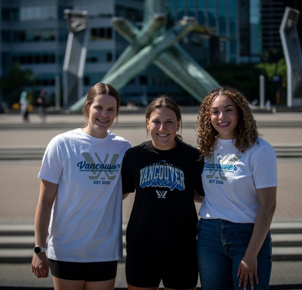 Three women, arm-in-arm, smile and look at the camera.