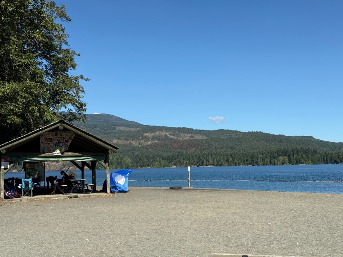 A beach shelter with two people inside, near a lake a small mountains. 