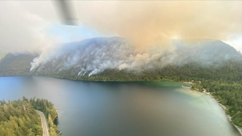 Orange smoke arises from a forested hill near a picturesque lake.