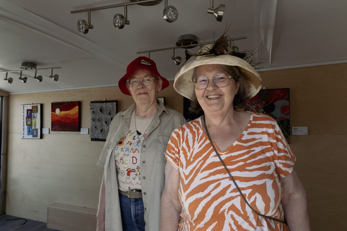 Two women stand in a small trailer with felt and wool art pieces hung behind them.