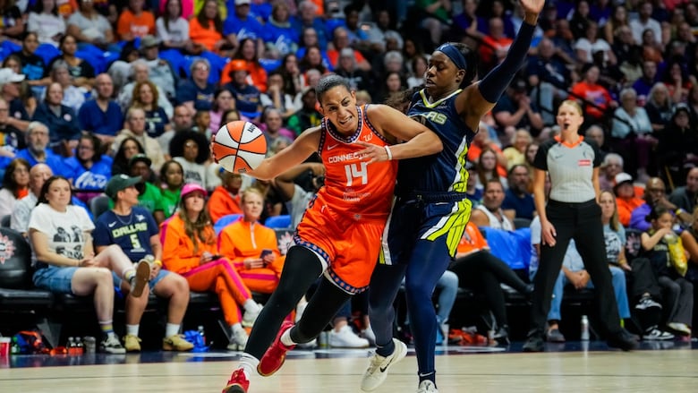 A female basketball player drives past an opponent with the ball in her right hand as fans watch from the stands.