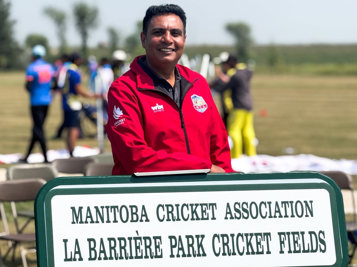 Man wears a red windbreaker jacket and stands behind a green and white sign that says "Manitoba Cricket Association La Barrière Park Cricket Fields"