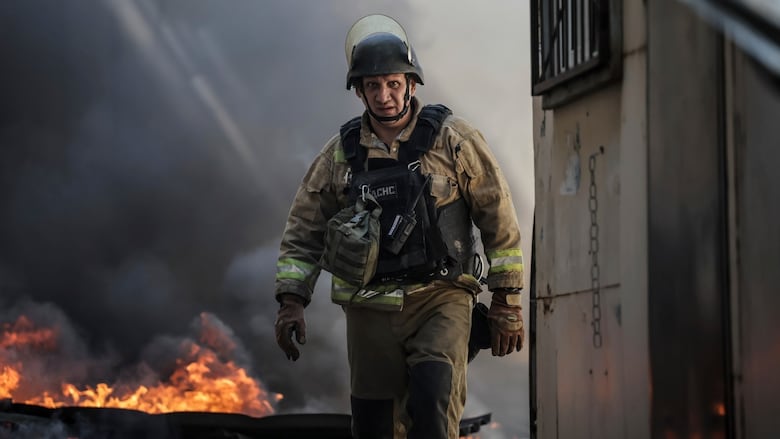 A man in firefighting equipment and a helmet walks toward the camera as a structure burns behind him. 