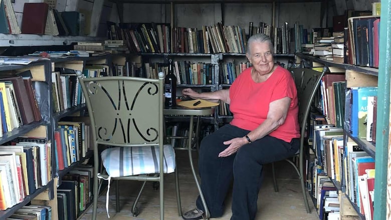 A white lady with white hair sitting in a shed surrounded by book shelves.