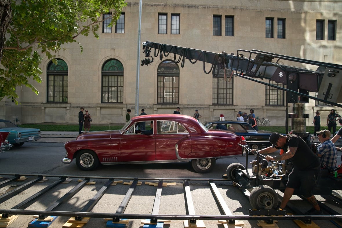 A red car is on a street, filmed by a filming team.