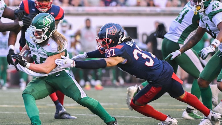 A football player in white and green holding a football evades an opponent in red and navy blue. 
