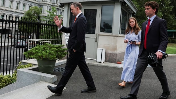 A man in a suit turns away from cameras as two younger people in a dress and a suit follow him. 