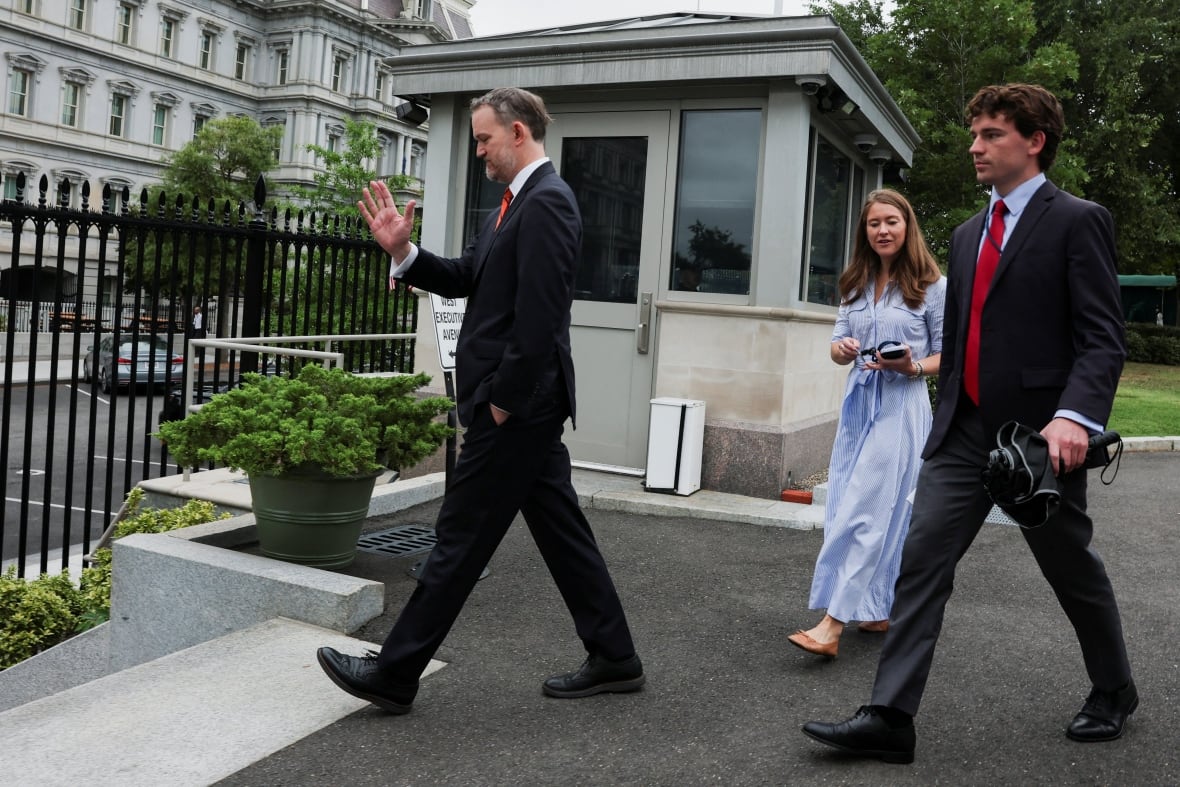 A man in a suit turns away from cameras as two younger people in a dress and a suit follow him. 