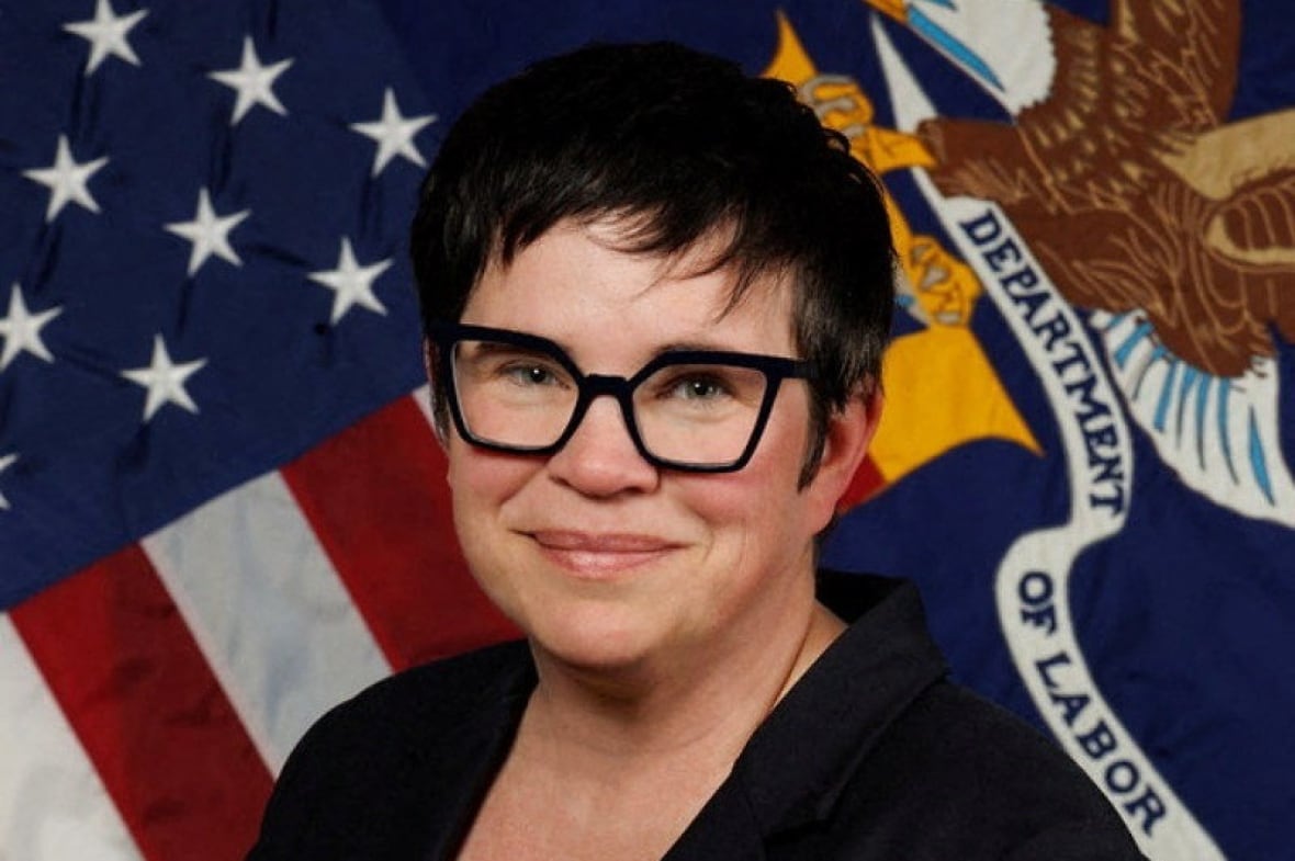 A woman with short black hair and glasses sitting in front of the American flag.