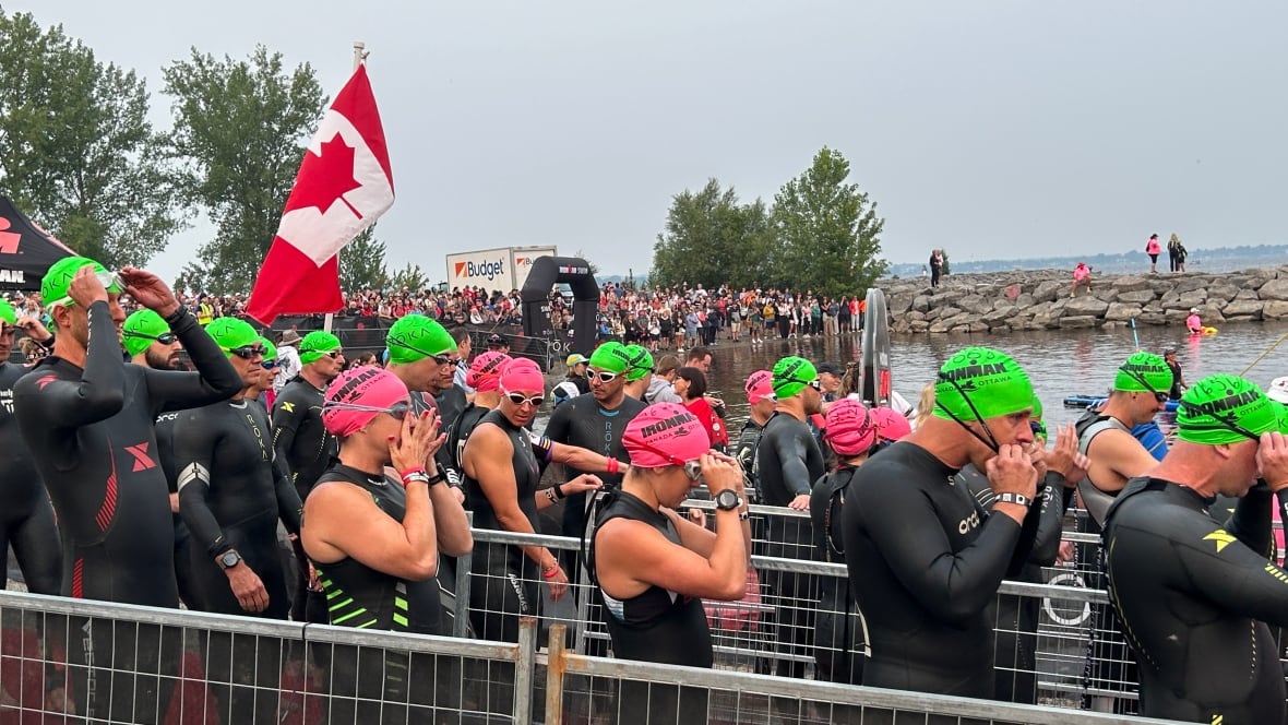 Dozens of athletes in swimming gear stand together in front of a body of water.