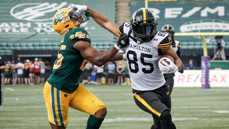 A Black football player wearing a white-and-black uniform is running down the field, with the ball craddled in his left arm. With his right arm, he is pushing an opposing player in a green-and-yellow uniform away from him.
