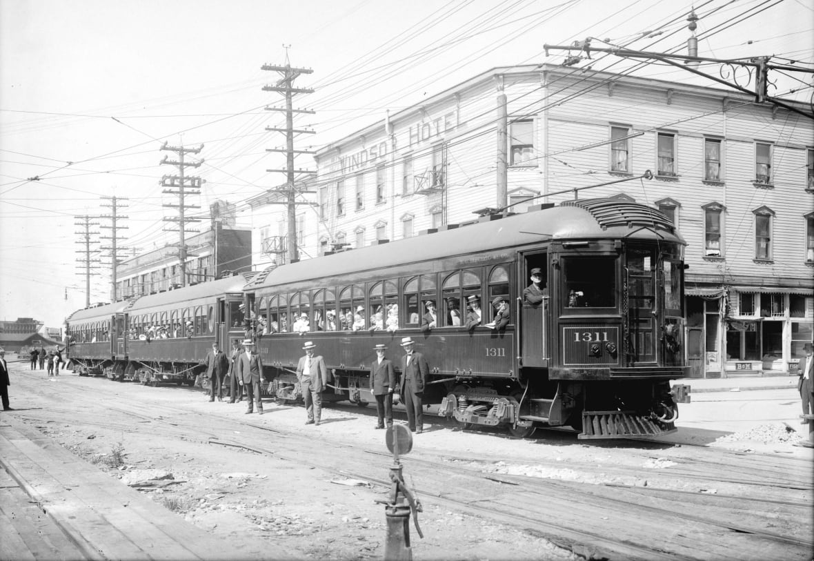 A black-and-white picture of a historical tram with people on it in a city centre.