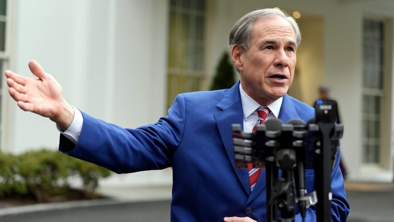 A man in a blue suit stands in front of an array of microphones and gestures while speaking. 