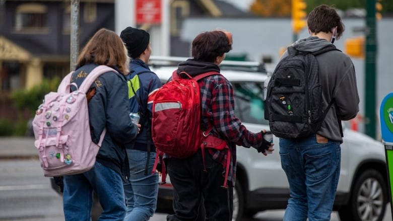 Teenagers are seen from behind as they walk down a street wearing backpacks.