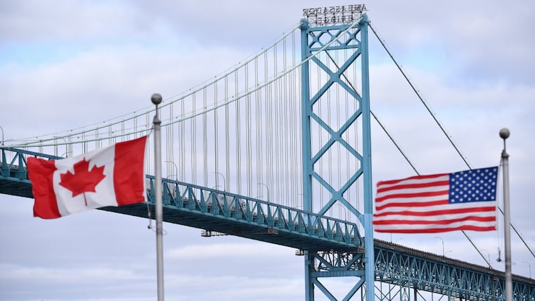 Canadian and American flags fly near the Ambassador Bridge at the Canada-USA border crossing in Windsor, Ont. On the left is the red and white Canadian flag, with a maple leaf in the centre and red vertical bands on the left and right sides. On the right is the American flag, with its red and white horizontal stripes and white stars on a blue background. The bridge spans across the image in the background against an overcast cloudy sky.