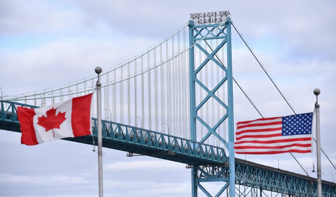 Las banderas canadienses y estadounidenses vuelan cerca del Puente del Embajador en el cruce fronterizo de Canadá-YUSA. A la izquierda está la bandera canadiense roja y blanca, hay bandas verticales rojas en la hoja de arce media y los lados izquierdo y derecho. A la derecha hay una bandera estadounidense a la derecha, con sus rayas horizontales rojas y blancas y estrellas blancas en el fondo. El puente se extiende por toda la imagen contra el cielo nublado.