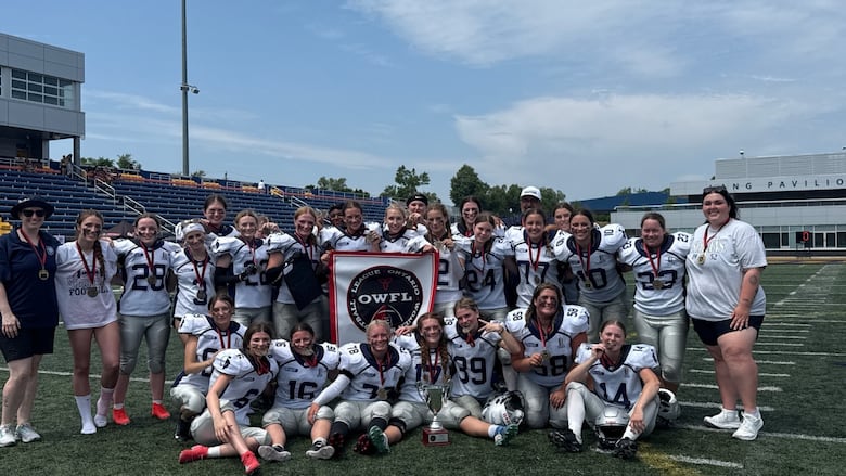 A group of female football players in white jerseys pose for a photo and hold a banner ono a football field.