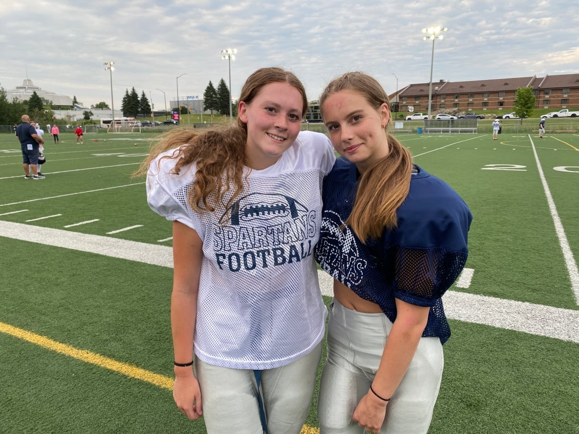 Two young girls in football jerseys stand on a football field and smile.