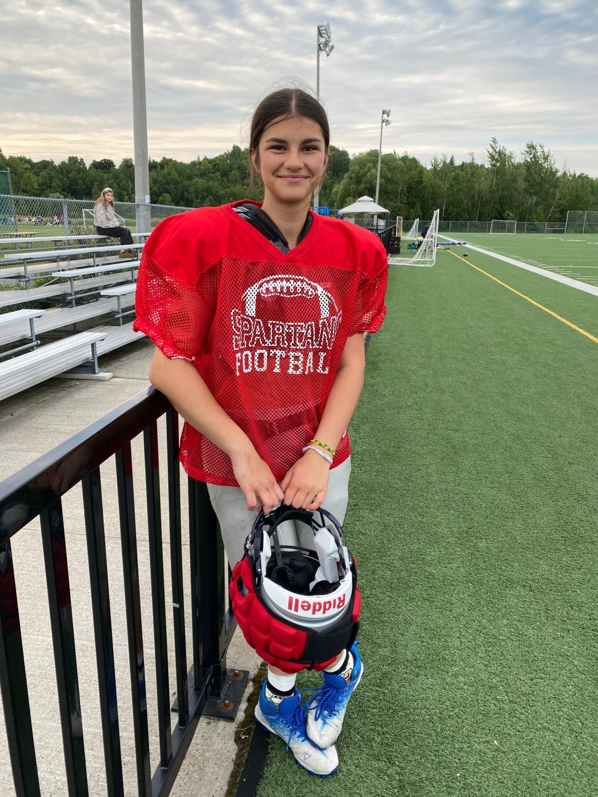 A teenaged girl in a red football jersey holds a football helmet on a football field.