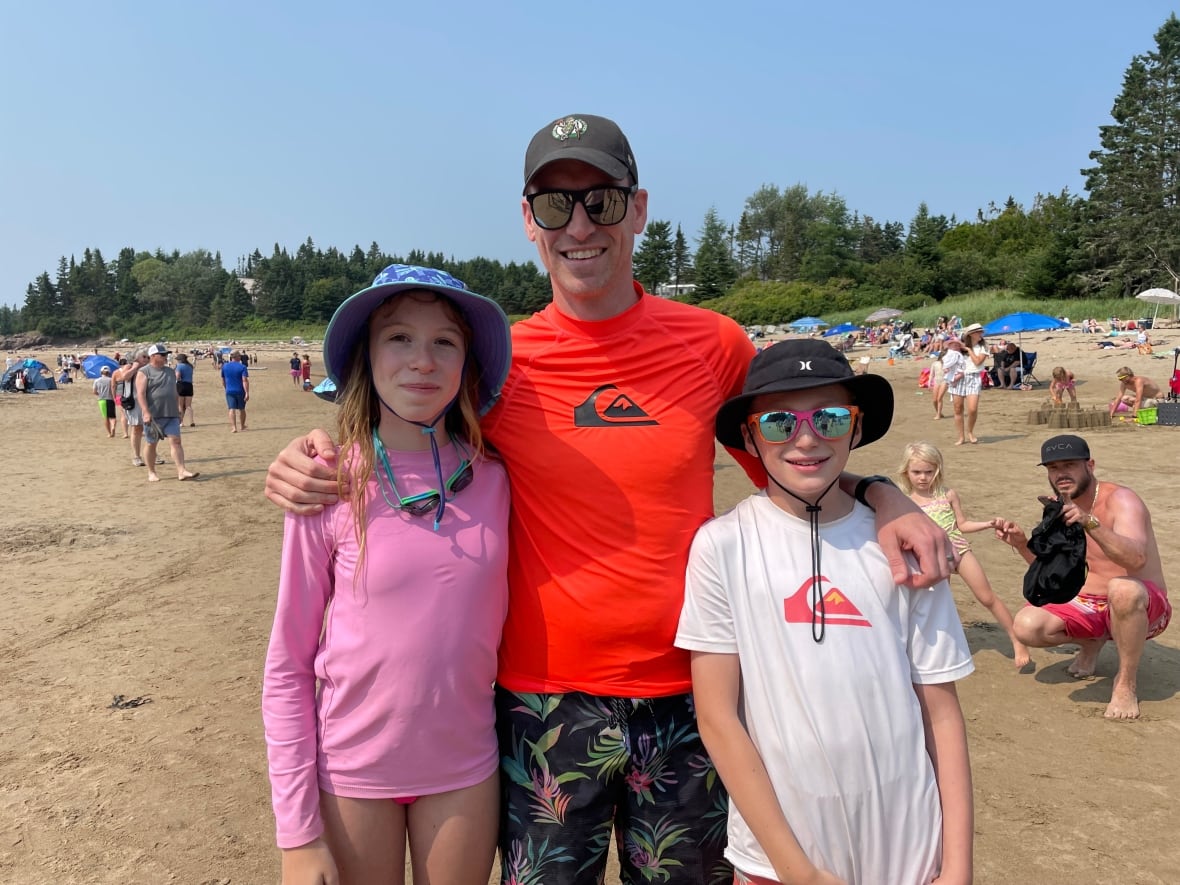 A father with his two kids on a sandy beach with a crown of people and sand sculptures.