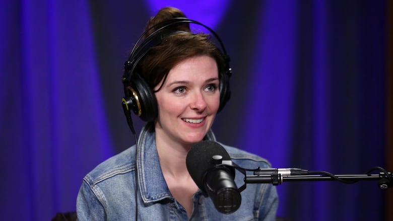 A smiling woman wearing over-ear headphones sits in front of a studio microphone.