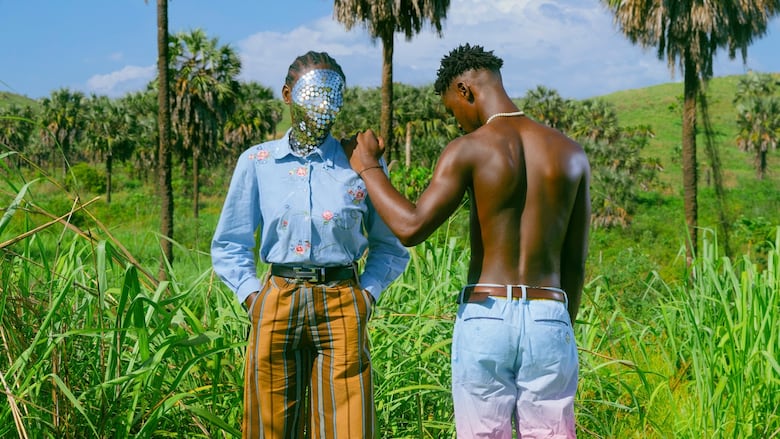 Standing in a grassy field with palm trees, one person wears a mirrored mask, striped pants and a patterned button-up, while another person wearing jeans without a top leans against them, facing away from the camera.