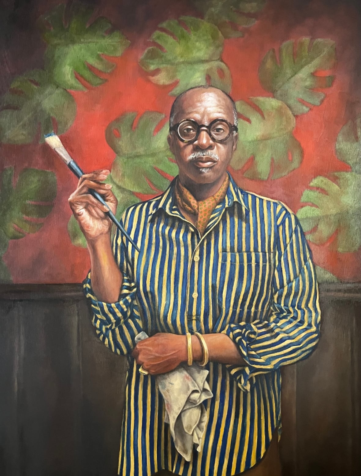 The painted portrait shows a man wearing round glasses, a blue-and-yellow striped shirt and an ascot. He is holding a paintbrush in one hand.