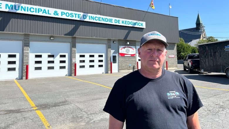 A man, straight-faced, wearing a ball cap, standing in front of a fire department