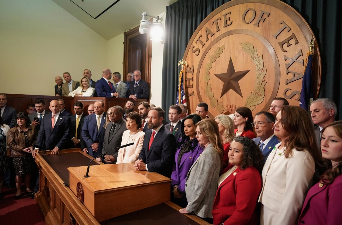 A large group of people stand behind a platform in a legislative building in front of a large wooden mark it says "Texas."