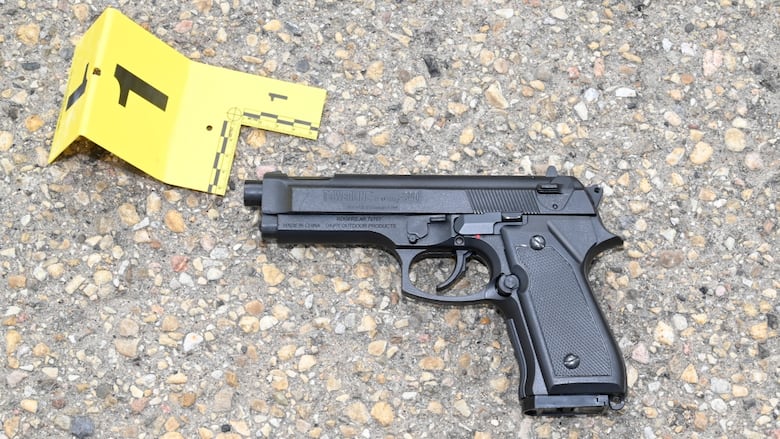 A realistic looking black BB gun sits on pavement. A yellow forensic marker sits next to the BB gun.