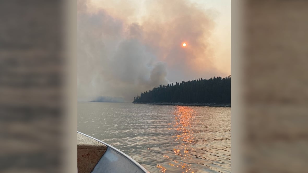 The front end of a boat can be see on the water as a fire burns a forest in the distance.